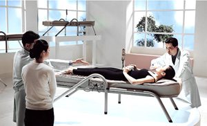 A woman is standing in front of two people laying on beds.